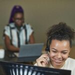 Smiling woman wearing a headset working at a call center desk with laptop.