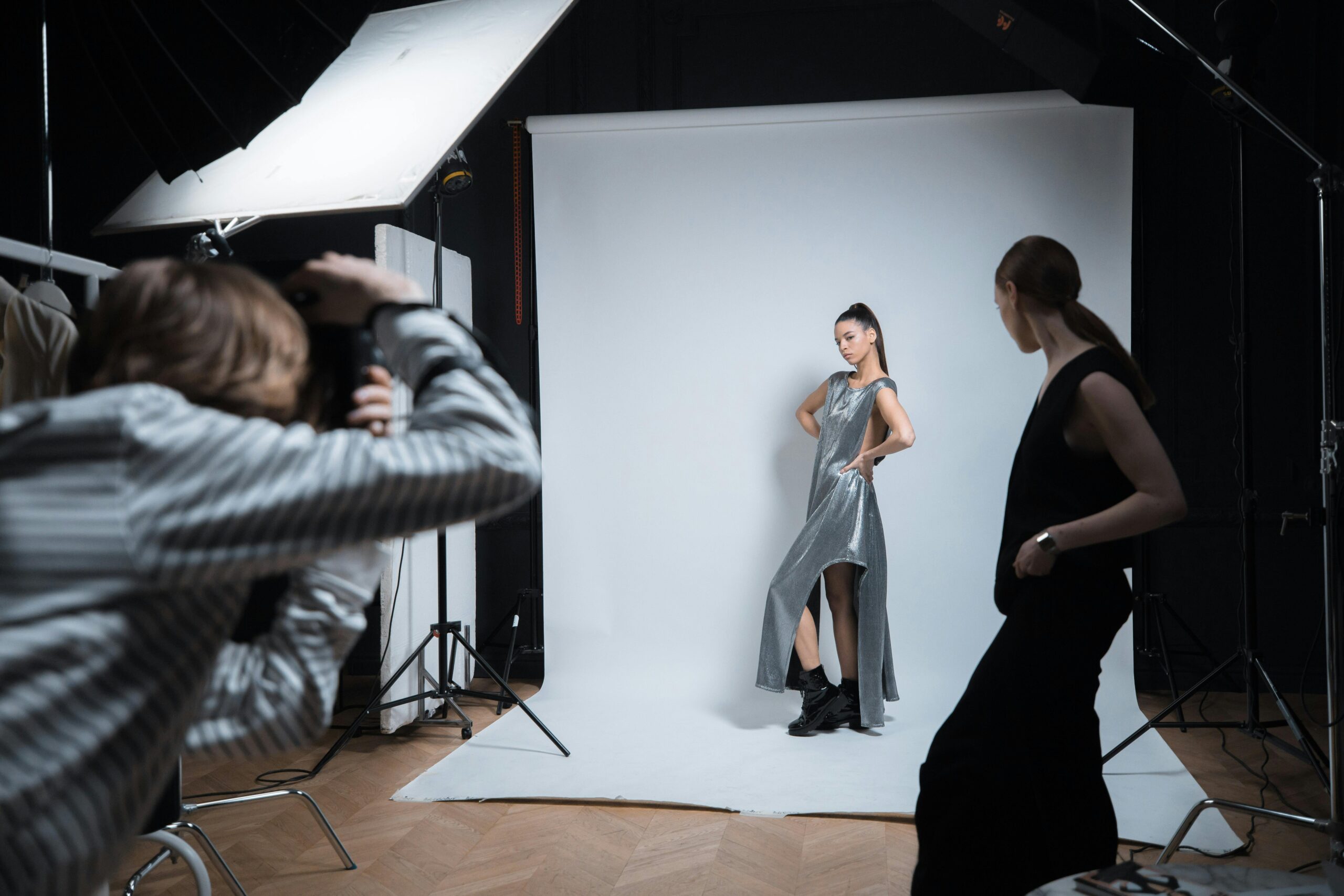 A professional studio shoot featuring a model in a silver dress with a photographer and assistant.